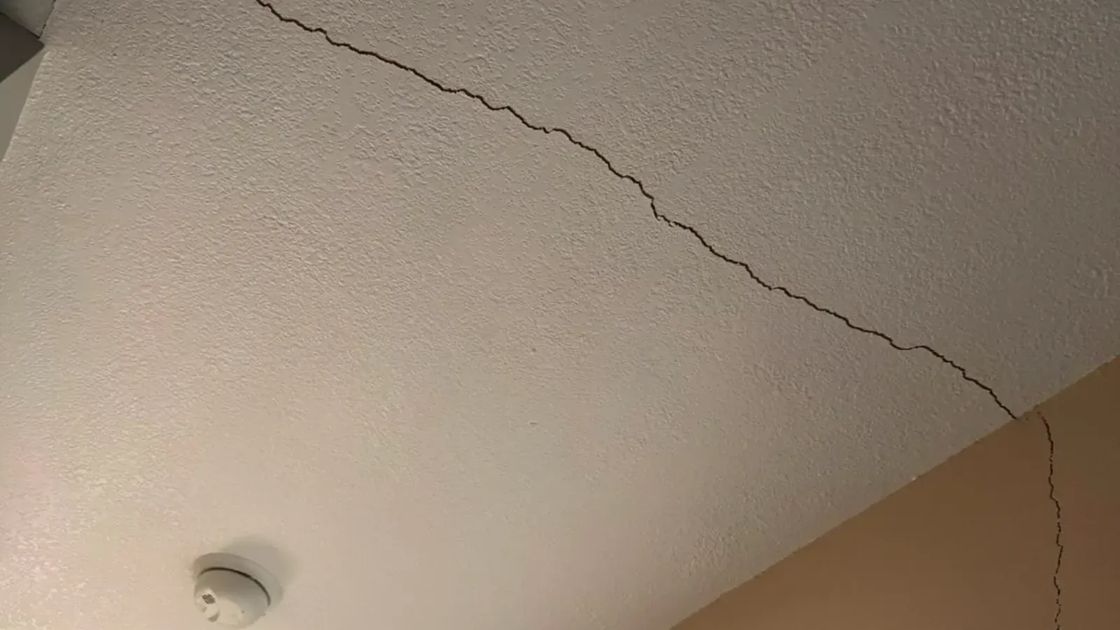 A long crack running diagonally across a lightly textured painted ceiling toward the corner where the ceiling meets the wall, with a round white smoke detector mounted nearby