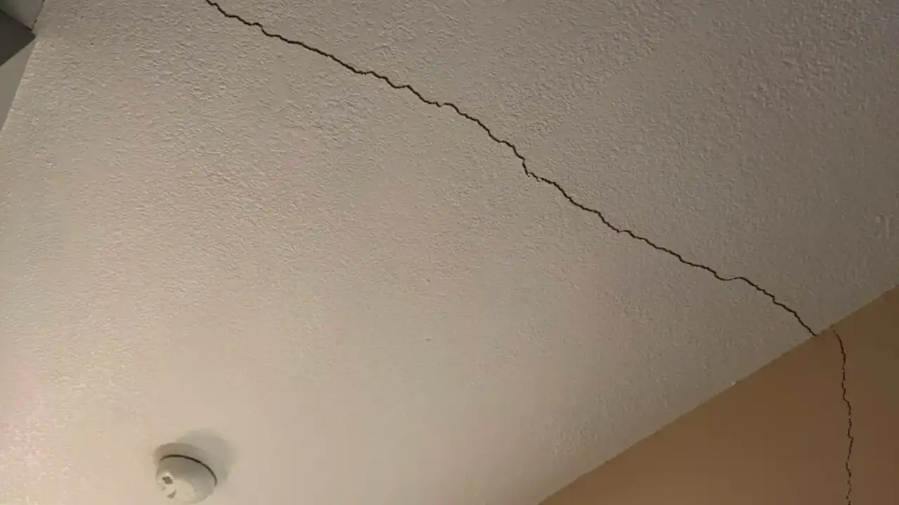 A long crack running diagonally across a lightly textured painted ceiling toward the corner where the ceiling meets the wall, with a round white smoke detector mounted nearby