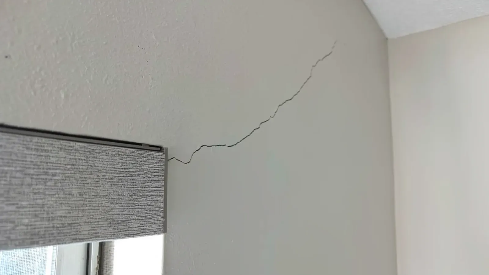 A hairline stress crack running diagonally across a painted interior wall from the head of a window casing up toward the wall-and-ceiling corner