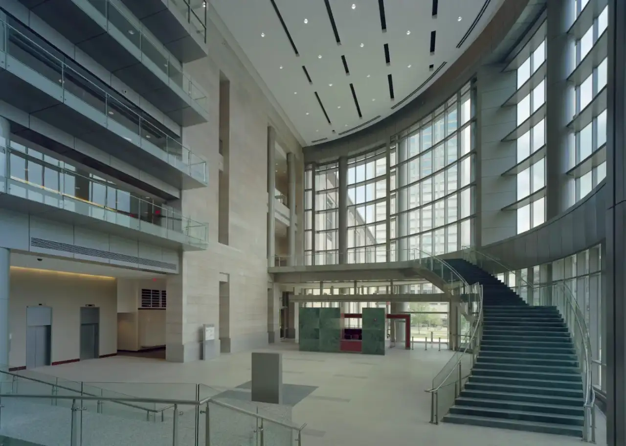 Soaring civic-building atrium in Springfield — a multi-story glass curtain wall, a sweeping stone staircase, and stone-clad walls with smooth plaster-finished surfaces under a finished drywall ceiling