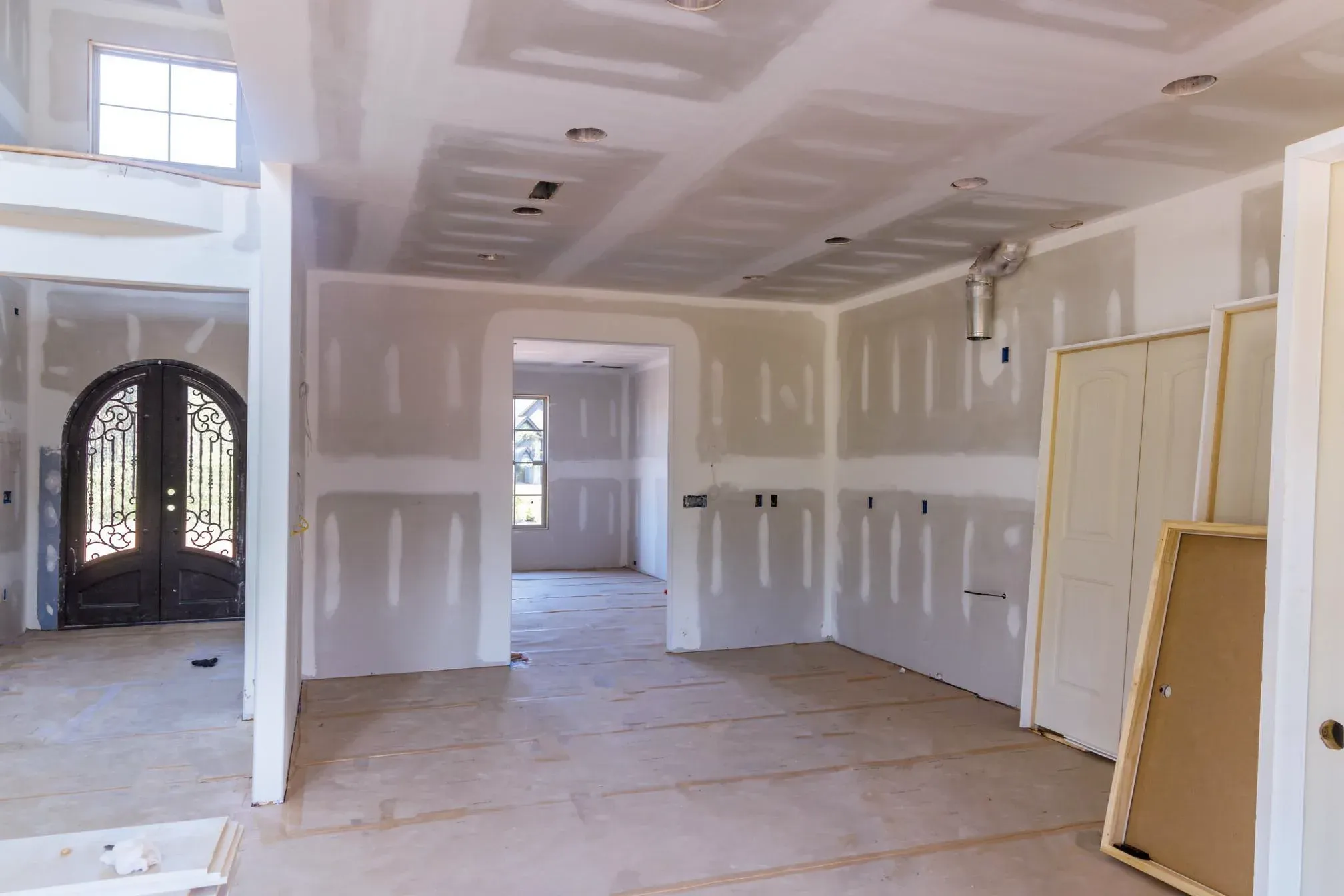 Open interior with taped and skim-coated drywall walls and ceiling, a wrought-iron front entry door, stacked interior doors, and a bare plywood floor